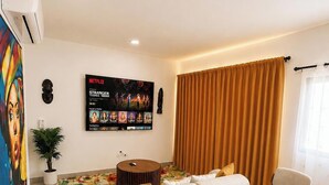 A living room with a sofa set, a dining table with chairs, a TV mounted on the wall, and a large window with curtains. - ANYA 6 Downtown Bayahíbe Retreat Pool  (Bayahíbe)