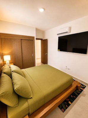 Un dormitorio con una cama, una mesita de noche, una lámpara, un televisor montado en la pared y un equipo de aire acondicionado.