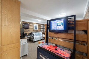 Une chambre d’hôtel équipée d’un lit, d’un bureau avec un ordinateur, d’un baby-foot et d’un téléviseur diffusant un match.