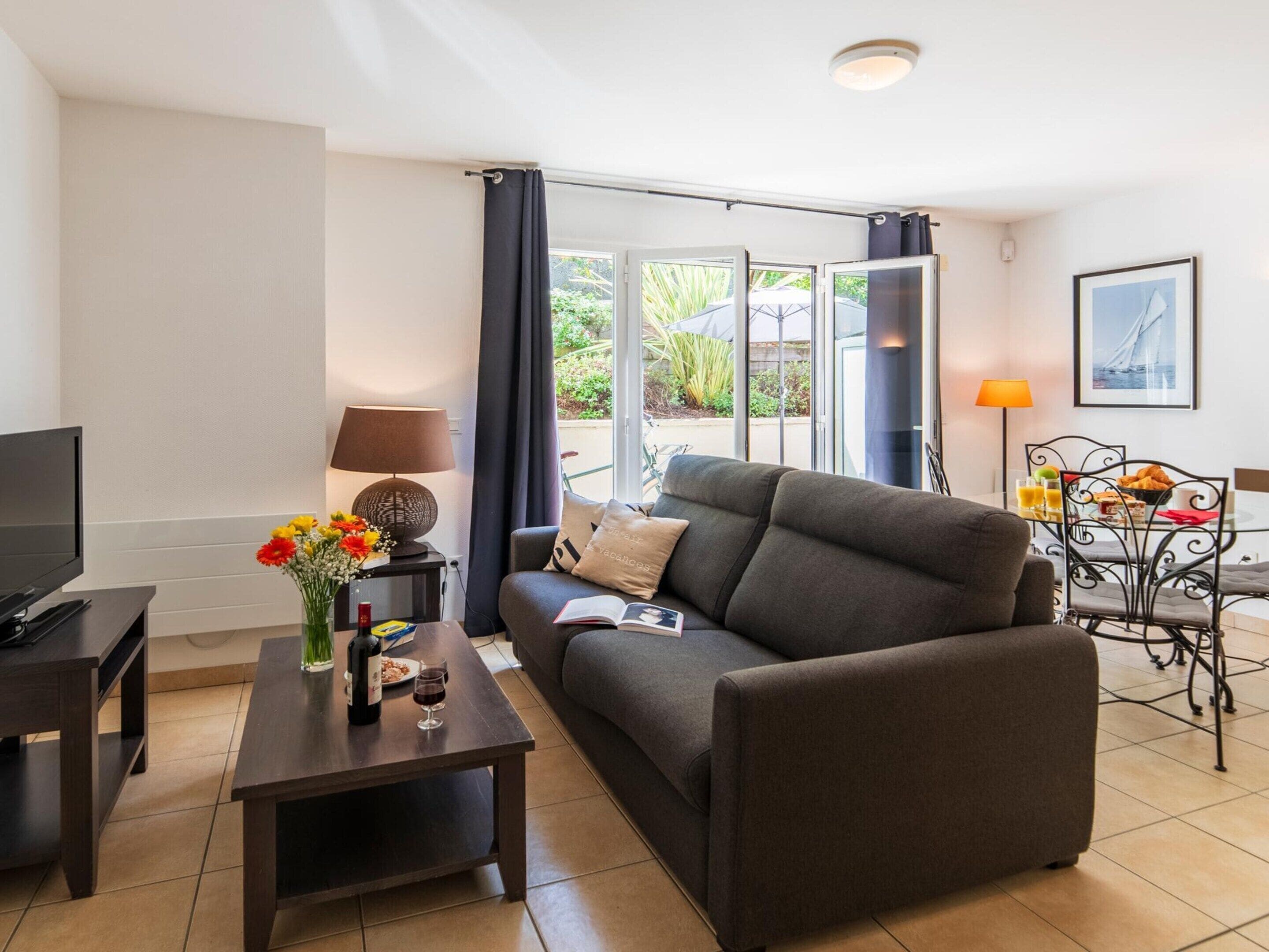 A modern living room with a sofa, a coffee table with a vase of flowers, a television, and a dining area with a table set for breakfast.