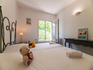 Habitación de hotel con dos camas, una mesita de noche con lámpara, un osito de peluche, un cartel y vistas a zonas verdes por la ventana.