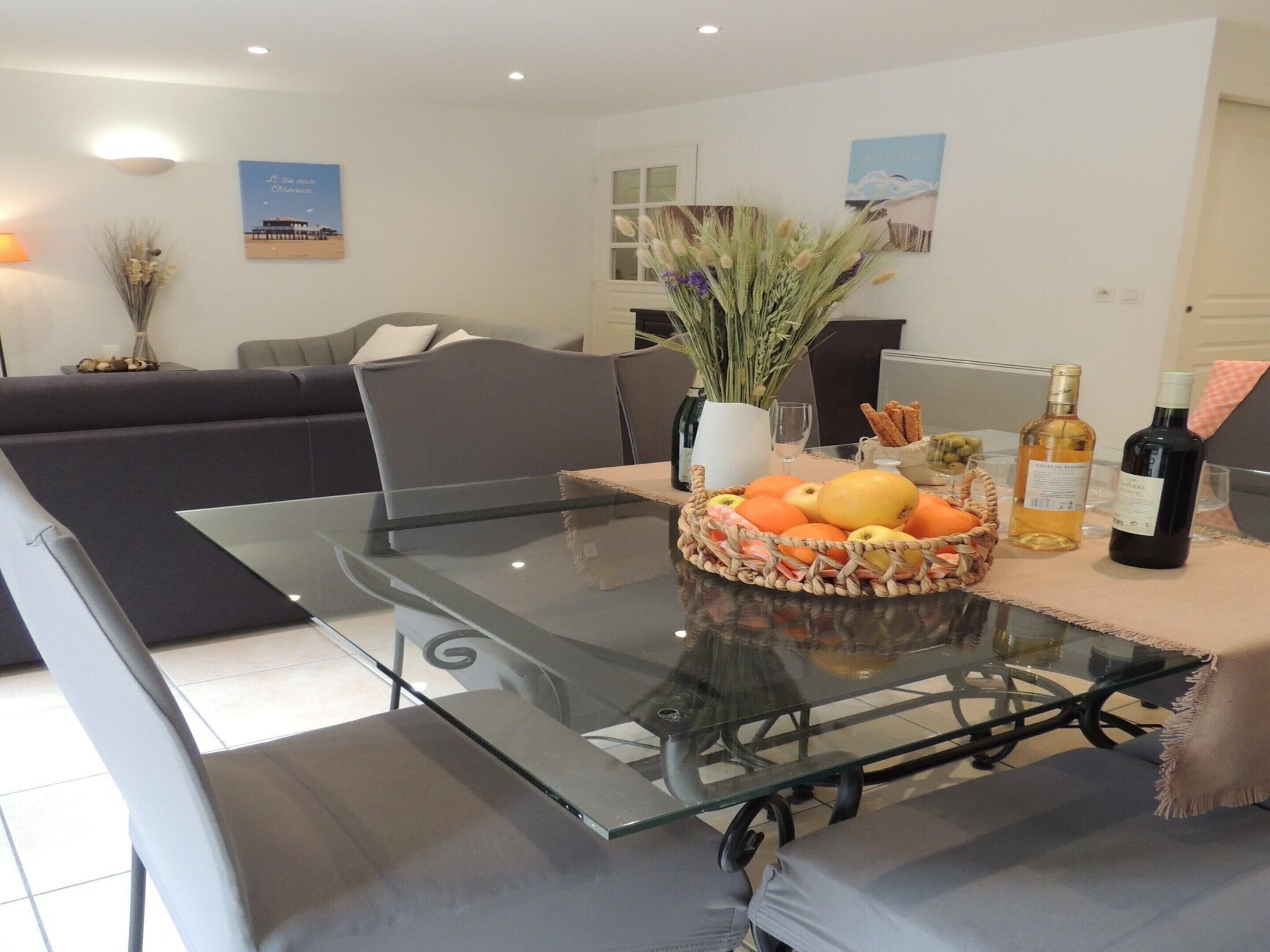 A modern living room with a glass-top dining table, grey sofas, and a decorative vase with dried flowers.