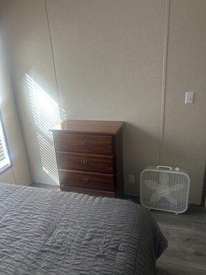 A bedroom with a bed, a wooden dresser, a white fan, and a window with blinds.
