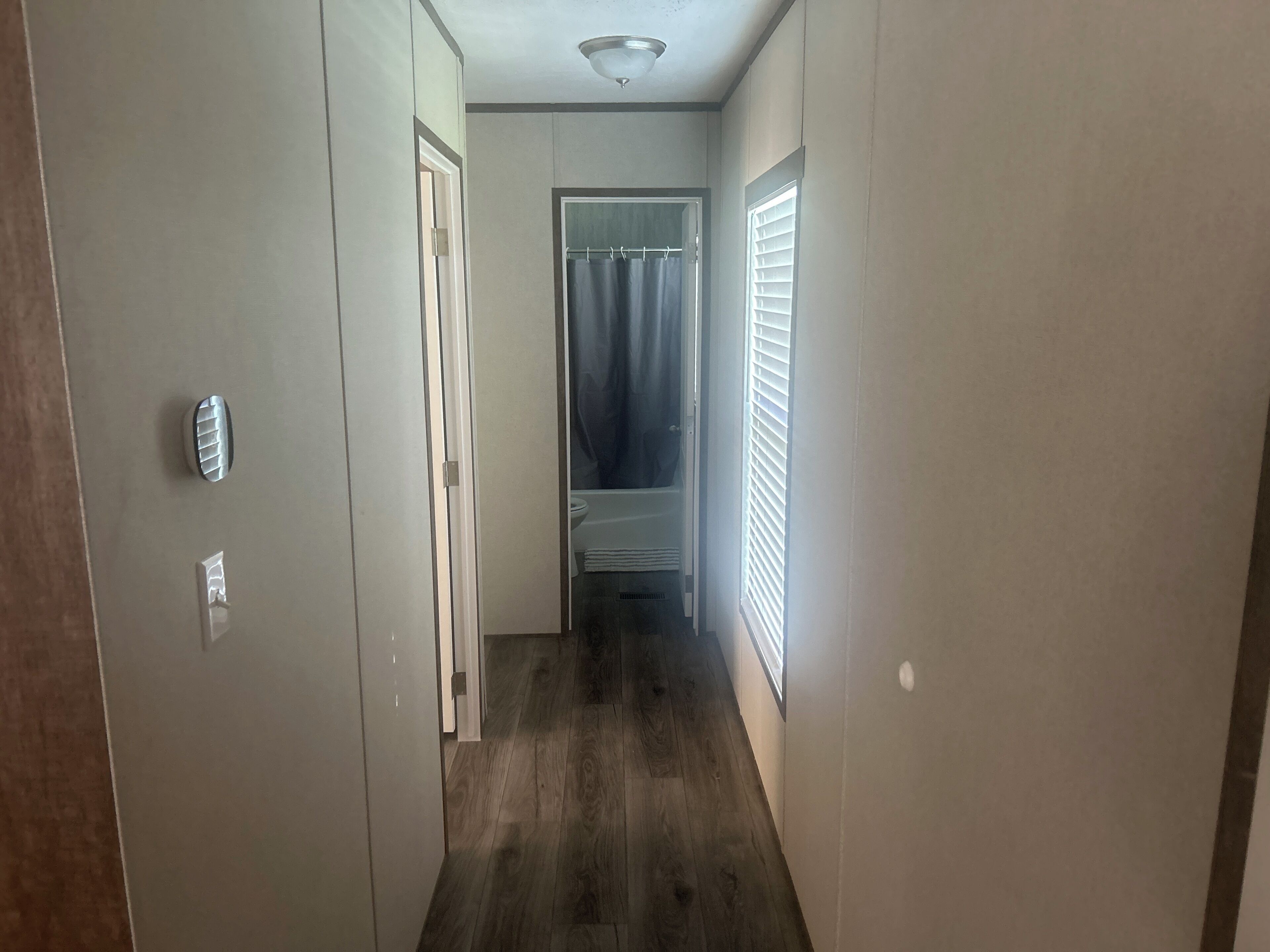 A narrow hallway with a wooden floor, a door on the left, and a window with blinds on the right.