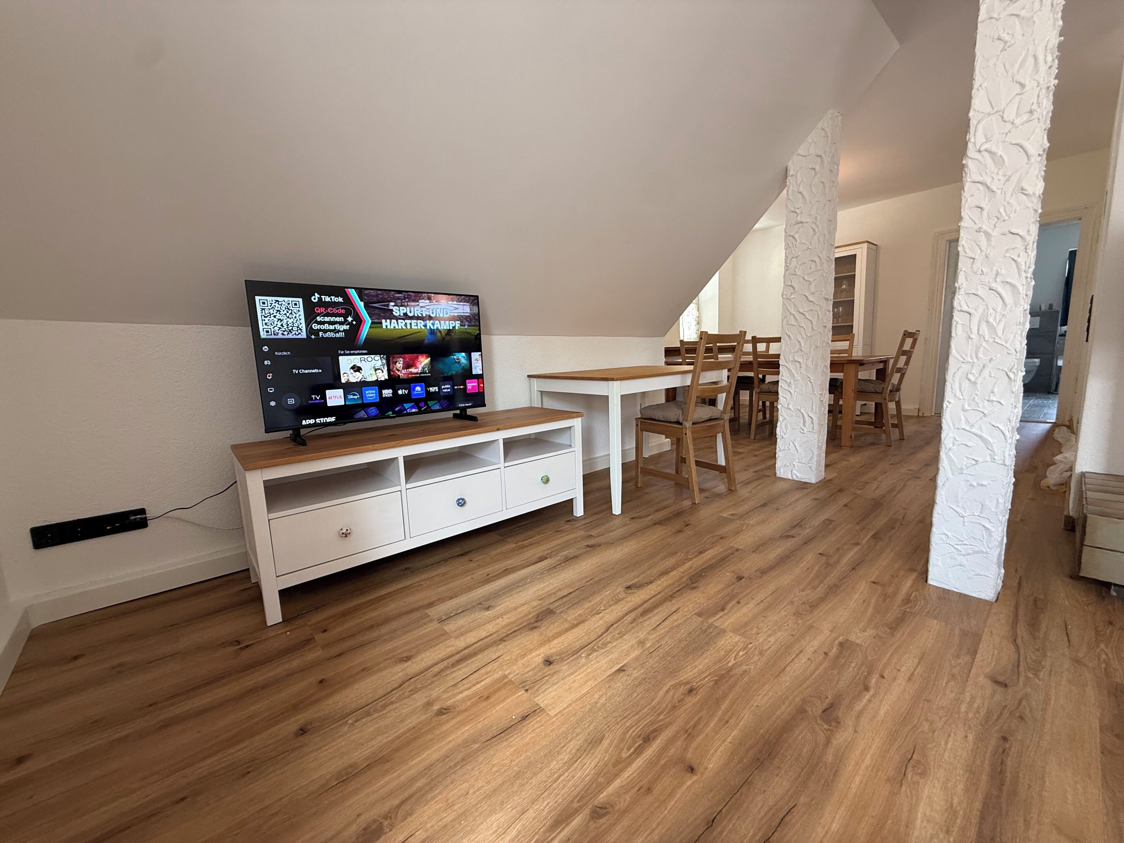 Ein modernes Wohnzimmer mit Flachbildfernseher, Holzboden und Essbereich mit Stühlen und Tisch.