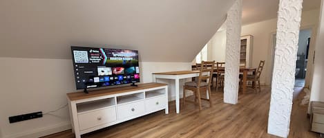 A modern living room with a flat-screen TV, wooden flooring, and a dining area with chairs and a table.