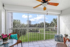 Ein Balkon mit Deckenventilator, eine Glastür zu einer grünen Fläche und ein Tisch mit Blumen.