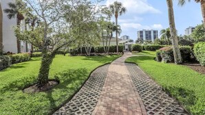 Caminho que serpenteia por um jardim bem cuidado, com palmeiras e sebes podadas.