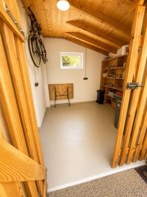 A room with a wooden ceiling, a window, a bicycle, shelves with various items, and a bench.