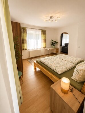 A bedroom with a bed, bedside table, desk, chair, and a plant.