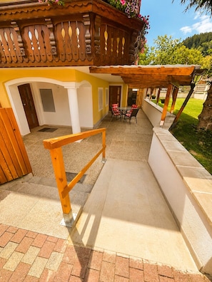 A covered outdoor seating area with a wooden pergola, a dining table with chairs, and a view of a lush garden.