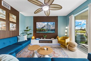 A modern living room with a blue sofa, two round coffee tables, a yellow armchair, a flat-screen TV mounted on a wooden panel, and a ceiling fan. - Luxury 3BR Resort Condo Near Disney - The Palms at Paradox (Kissimmee)