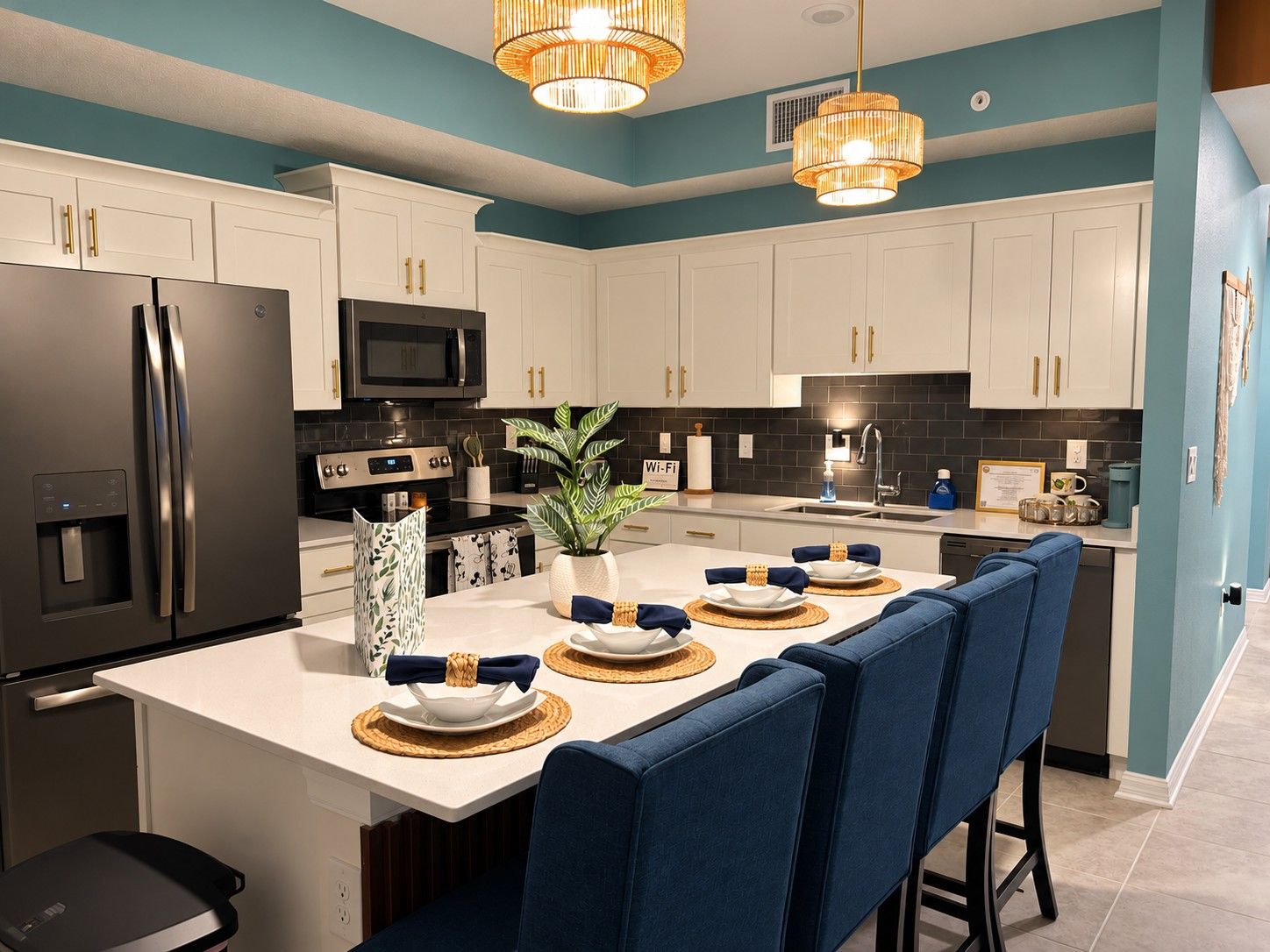 Cocina moderna con armarios blancos, refrigerador de acero inoxidable, microondas y pileta. Cuenta con un comedor con taburetes azules y una isla central con una planta y objetos decorativos.