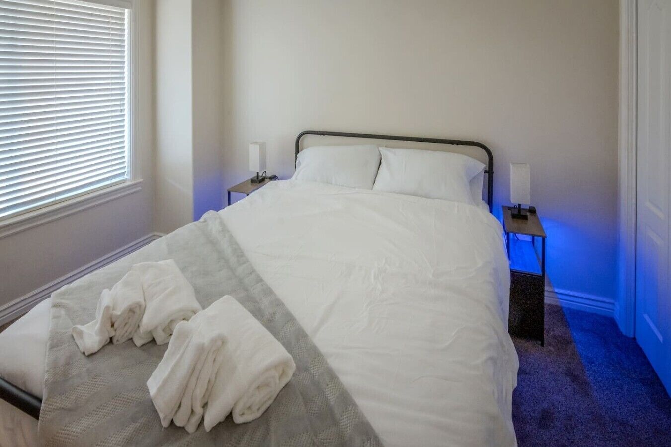 A neatly made bed with white linens and a folded towel on the side, a bedside table with a lamp, and a window with blinds.