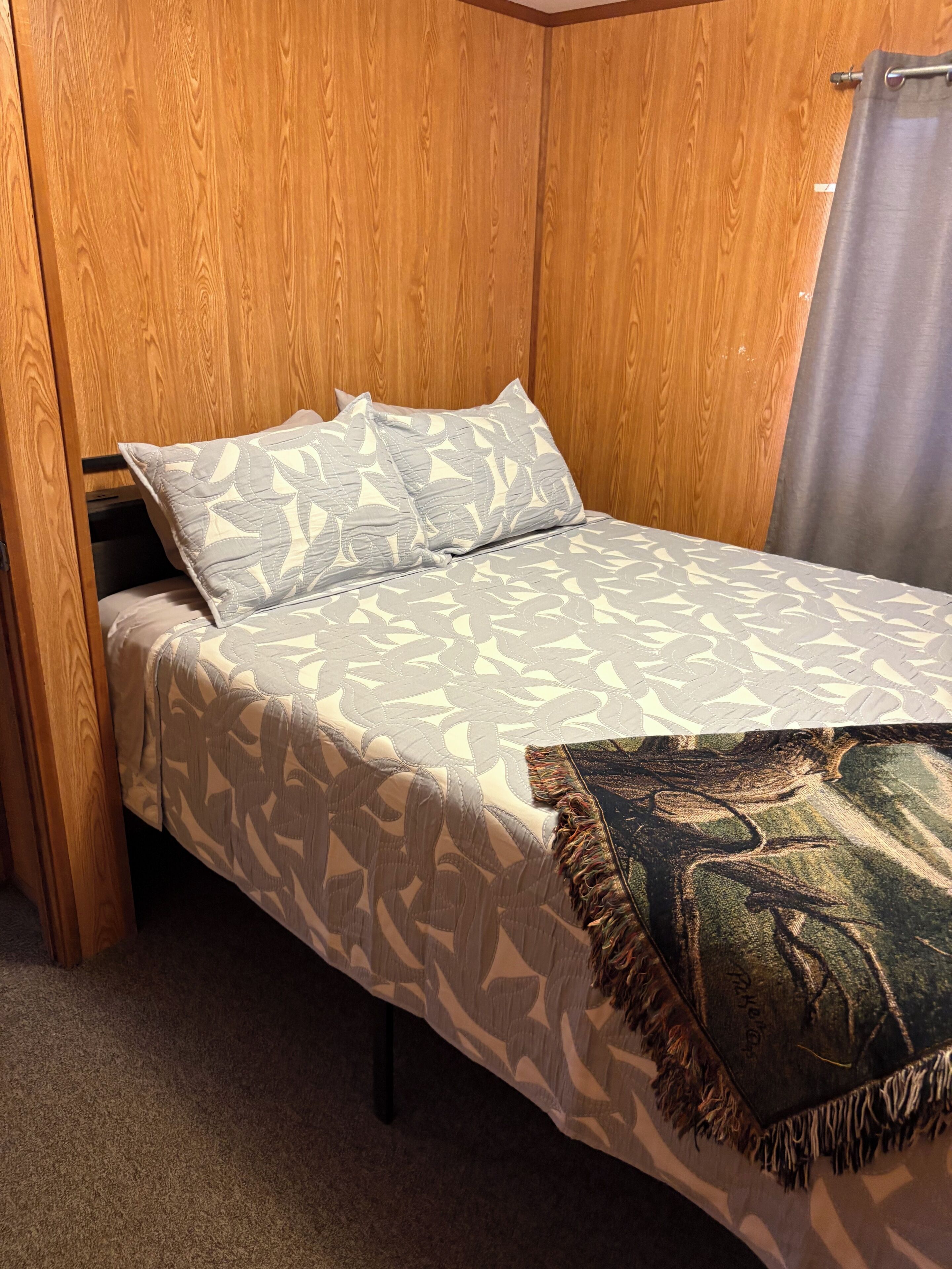 A single bed with a patterned bedspread and pillows, a wooden headboard, and a curtain on the window.