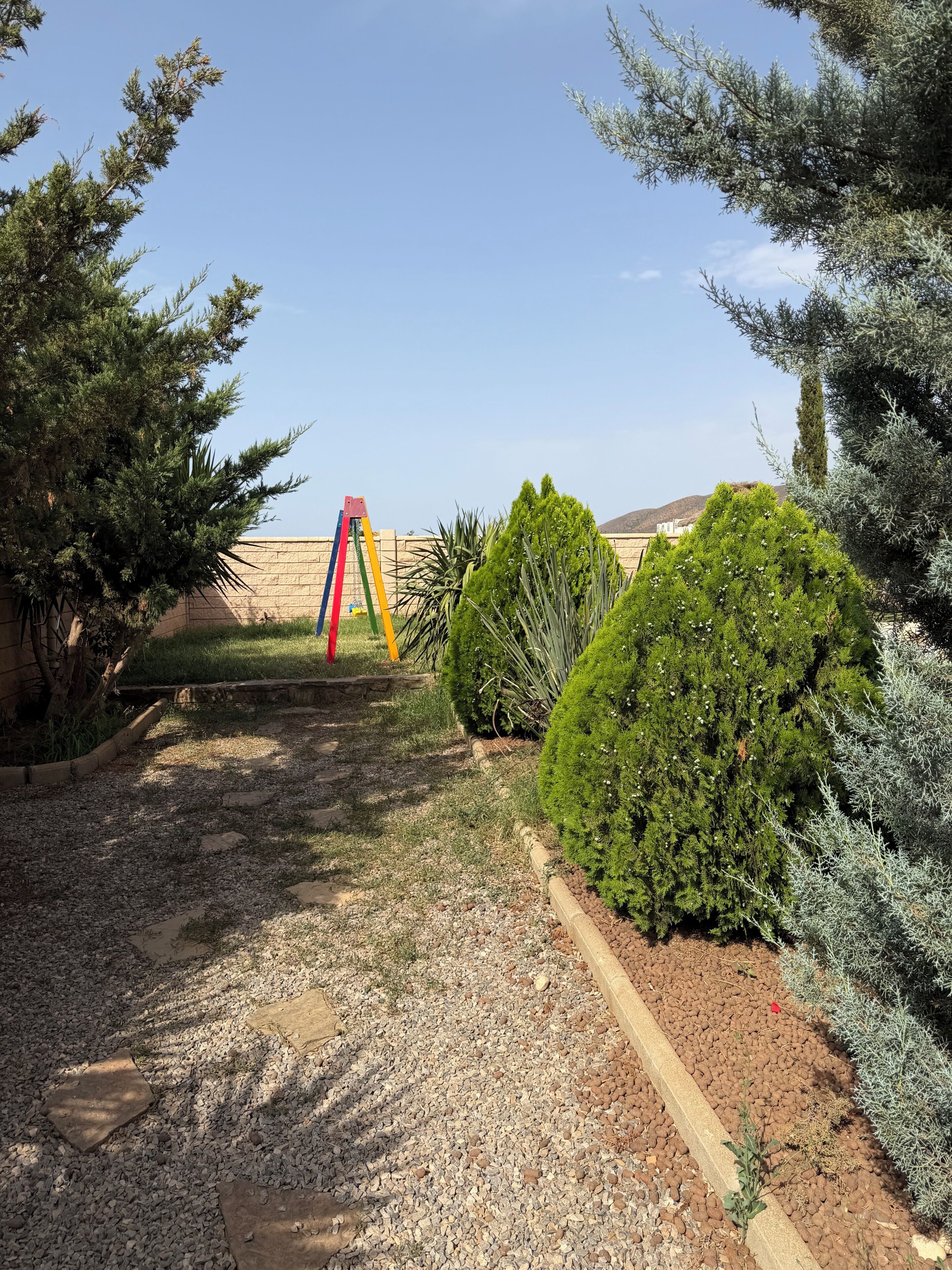 A garden with a gravel path, a colorful swing set, and various shrubs.