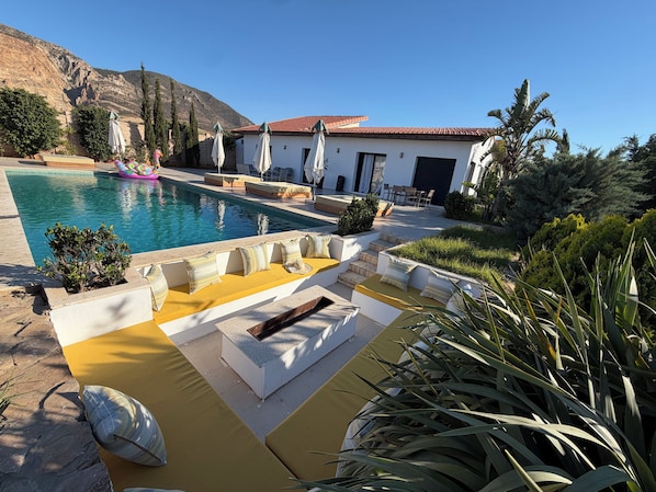 A pool area with a white building, outdoor seating, and a mountain backdrop.