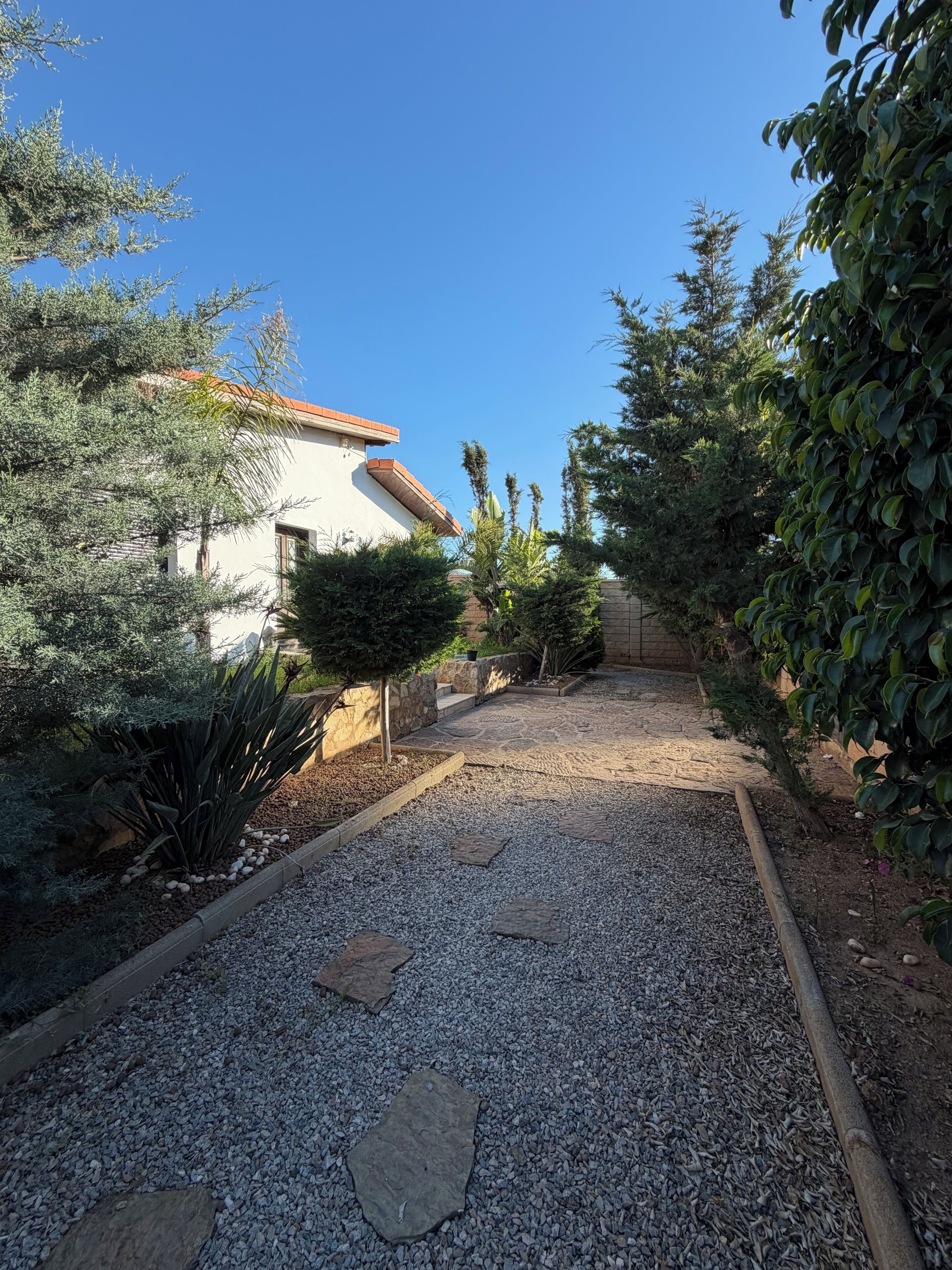 Un chemin de gravier mène à un bâtiment doté d’un toit en tuiles, qui est entouré de divers arbustes et plantes.