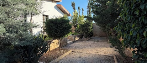 A gravel pathway leading to a building with a tiled roof, surrounded by various shrubs and plants.