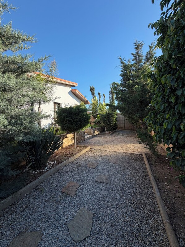 A gravel pathway leading to a building with a tiled roof, surrounded by various shrubs and plants.