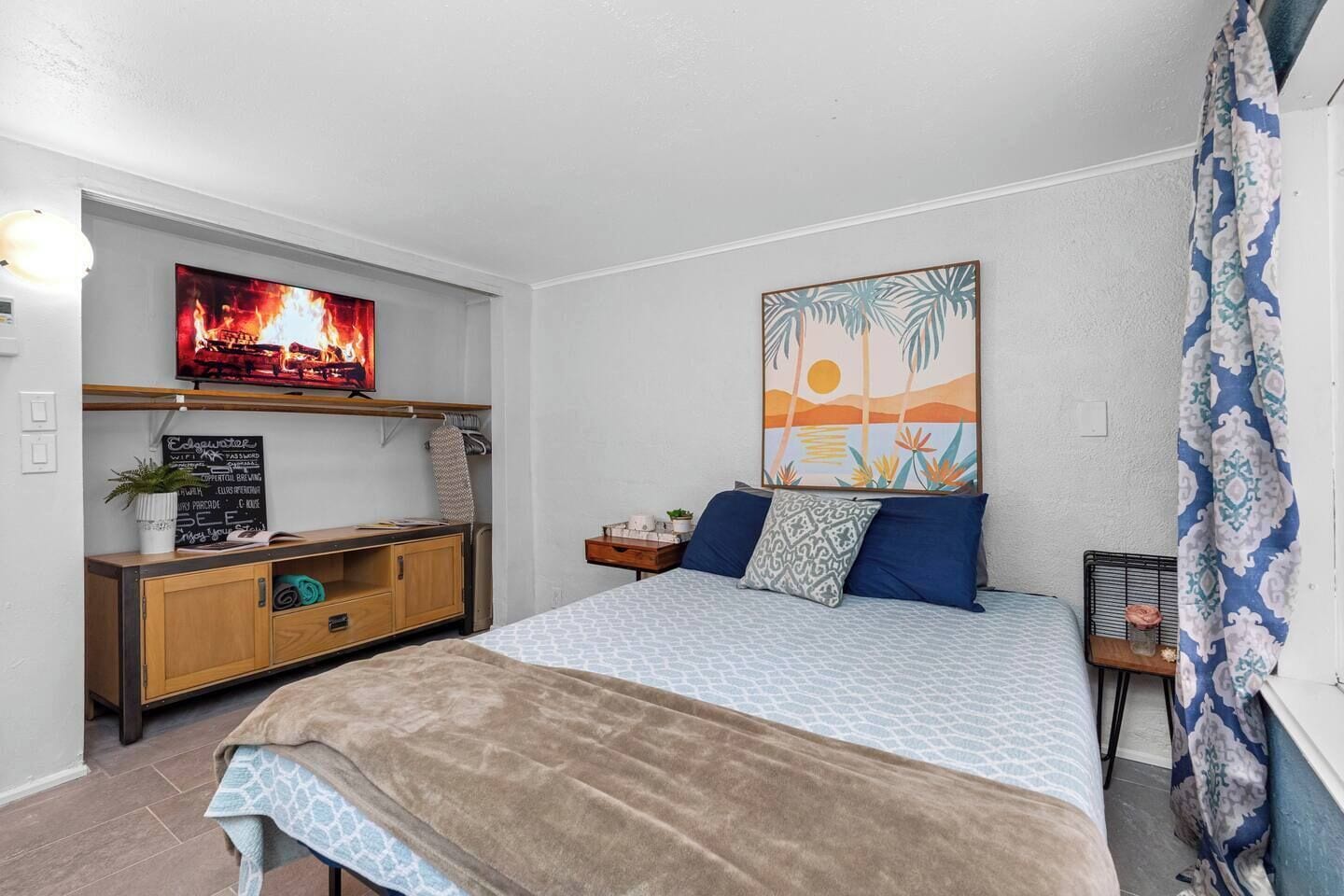A bedroom with a bed, a TV mounted on the wall, a desk with a chalkboard, and a window with curtains.