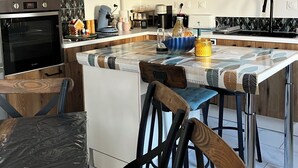A modern kitchen with wooden cabinets, a built-in oven, a coffee machine, and a dining area with a table and chairs. - Small vacation rental near the beaches (Triaize)