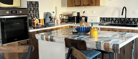 A modern kitchen with wooden cabinets, a built-in oven, a coffee machine, and a dining area with a table and chairs.