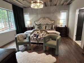A bedroom with a large bed, two nightstands, a chandelier, and a view of the outdoors. - USAFA Retreat, 4BR/ 4BA, Front Range Views, Nestled in the Pines (Monument)