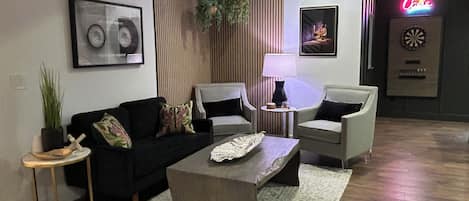 Un salon moderne avec un canapé noir, deux fauteuils, une table basse et une lampe. Un tableau encadré est accroché au mur, ainsi qu’une plante et un néon en arrière-plan.