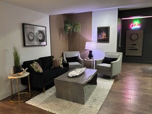 Ein modernes Wohnzimmer mit einem schwarzen Sofa, zwei Sesseln, einem Couchtisch und einer Lampe. An der Wand hängt ein gerahmtes Bild, eine Pflanze steht dort, und im Hintergrund ist eine Neonreklame zu sehen.