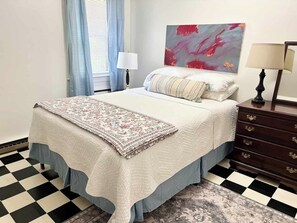 A bedroom with a bed, a nightstand, a lamp, a painting, and a patterned rug.
