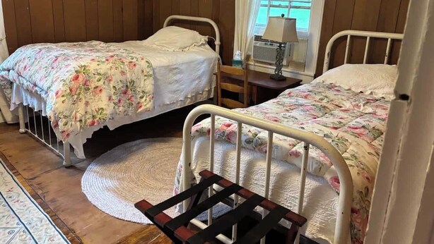 A room with two single beds, a wooden floor, a round rug, a desk with a lamp, and a window with curtains. - Farmville Lake House minutes from H-SC and Longwood (Farmville)