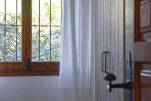 Eine Holztür mit Metallbeschlägen, ein Fenster mit einem Metallgitter und ein weißer Vorhang, der teilweise heruntergezogen ist.