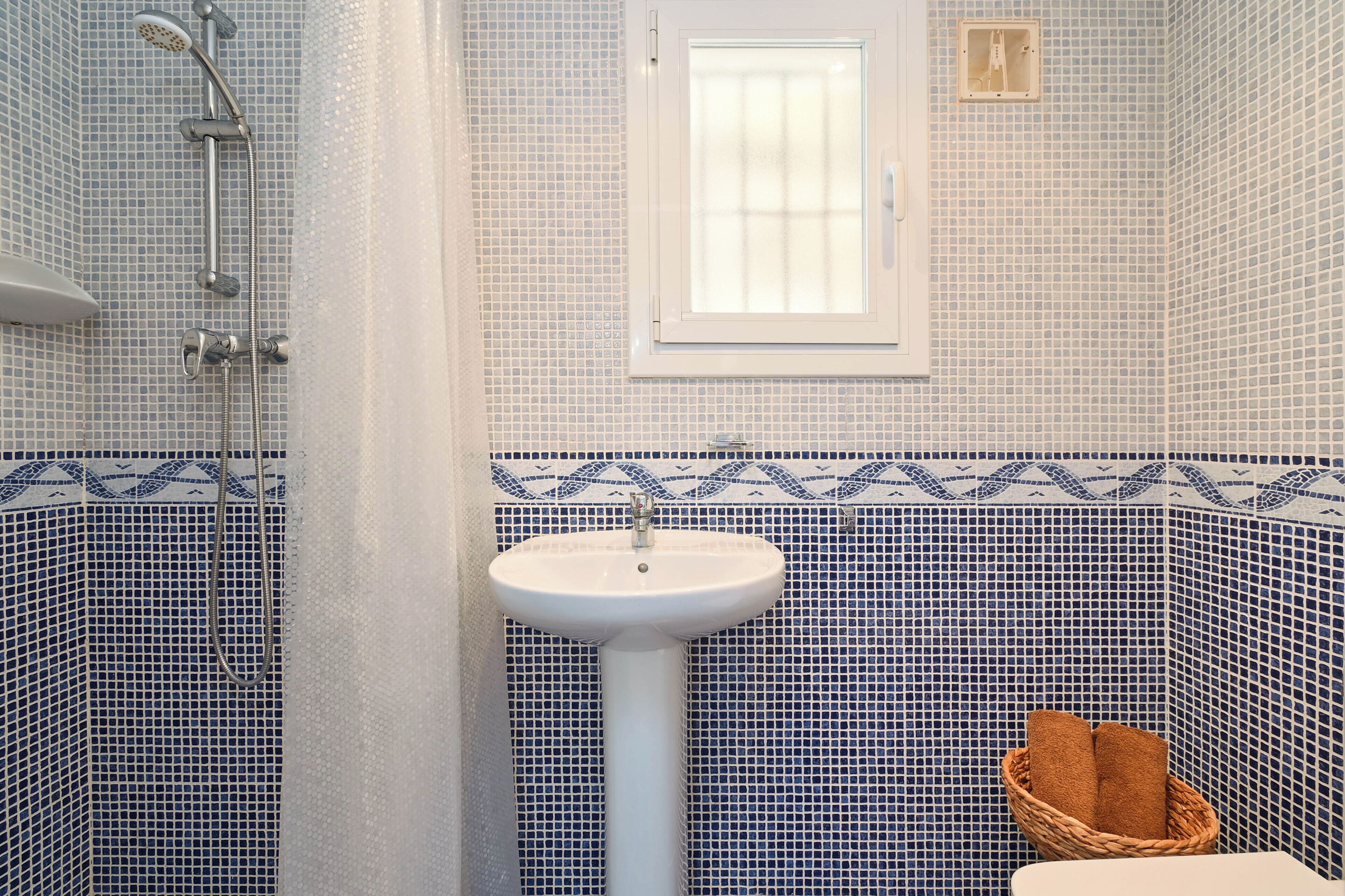 Une salle de bain avec un lavabo sur colonne blanc, une douche équipée d’un pommeau de douche à main et une fenêtre au-dessus du lavabo.
