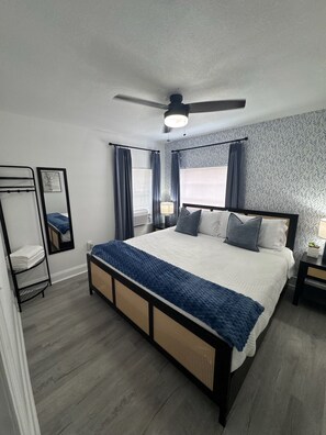 Une chambre moderne avec un lit, un ventilateur de plafond, une table de chevet avec une lampe, un miroir fixé au mur et une fenêtre avec des rideaux.