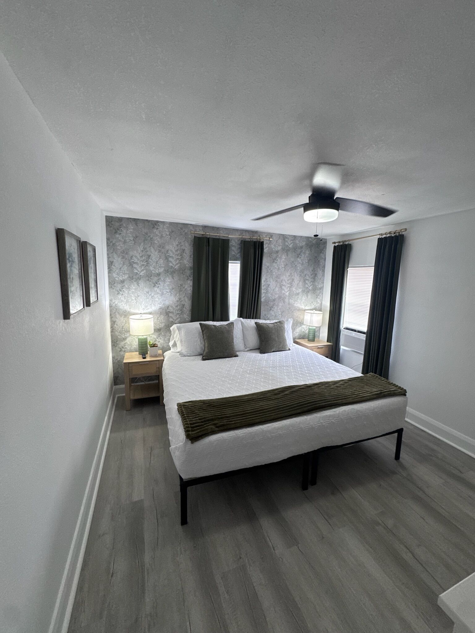 A bedroom with a large bed, two bedside tables with lamps, a ceiling fan, and a window with curtains.