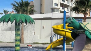 Un parc aquàtic amb un tobogan amb forma de dinosaure, una font d'aigua amb palmera i un edifici al fons.