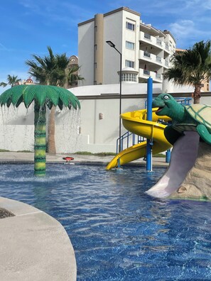 Ein Wasserpark mit einer Dinosaurier-Rutsche, einer Palme als Wasserspiel und einem Gebäude im Hintergrund.