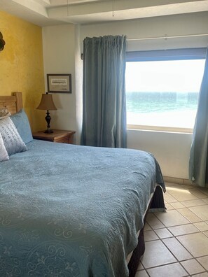 A bedroom with a bed, a nightstand, a lamp, a framed picture on the wall, and a window with curtains. - 1-bedroom Sandy Beach condo with ocean view and AC (Puerto Peñasco)