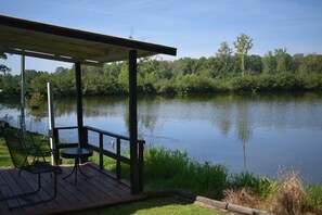 A wooden deck with a covered seating area overlooking a calm lake, surrounded by dense greenery. - Sunset Bay Waterfront Retreat (Springville)