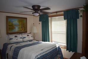 A bedroom with a bed, a ceiling fan, a painting, a window with curtains, and a lamp. - Sunset Bay Waterfront Retreat (Springville)