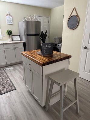A kitchen with a wooden countertop, a stainless steel refrigerator, a small island with a chalkboard sign, and a white door.