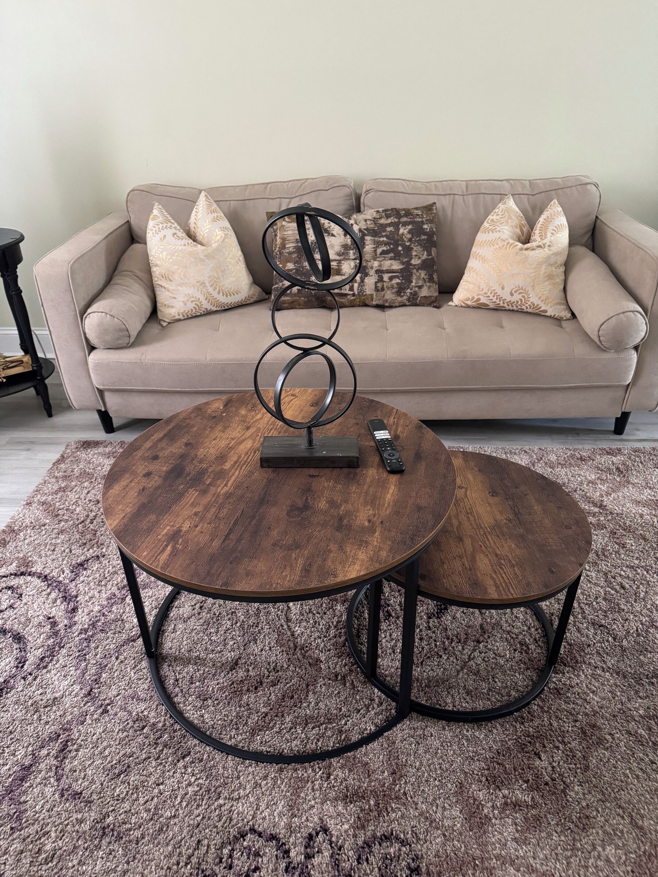 Ein Wohnzimmer mit einem beigen Sofa, gemusterten Kissen und einem runden Couchtisch mit Fernbedienung.