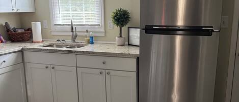 Cocina con armarios blancos, refrigerador de acero inoxidable, ventana con persianas, una pequeña planta en maceta y un letrero en la pared.