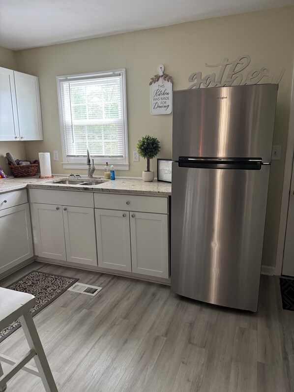 A kitchen with white cabinets, a stainless steel refrigerator, a window with blinds, a small potted plant, and a wall sign.
