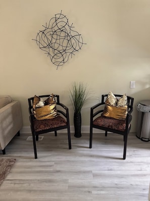 Two chairs with gold cushions, a decorative wall piece, and a potted plant in a room with light-colored flooring.