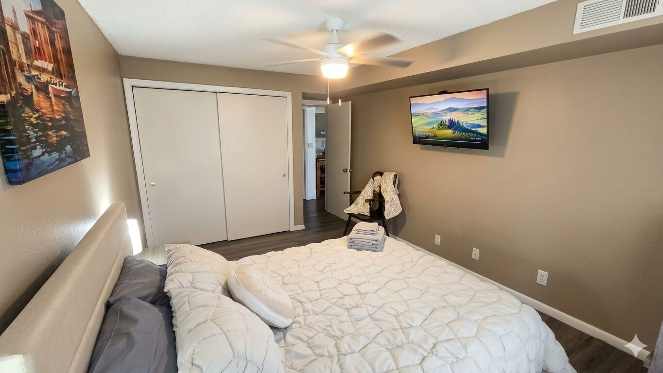 A bedroom with a bed, a ceiling fan, a TV on the wall, and a door leading to another room.