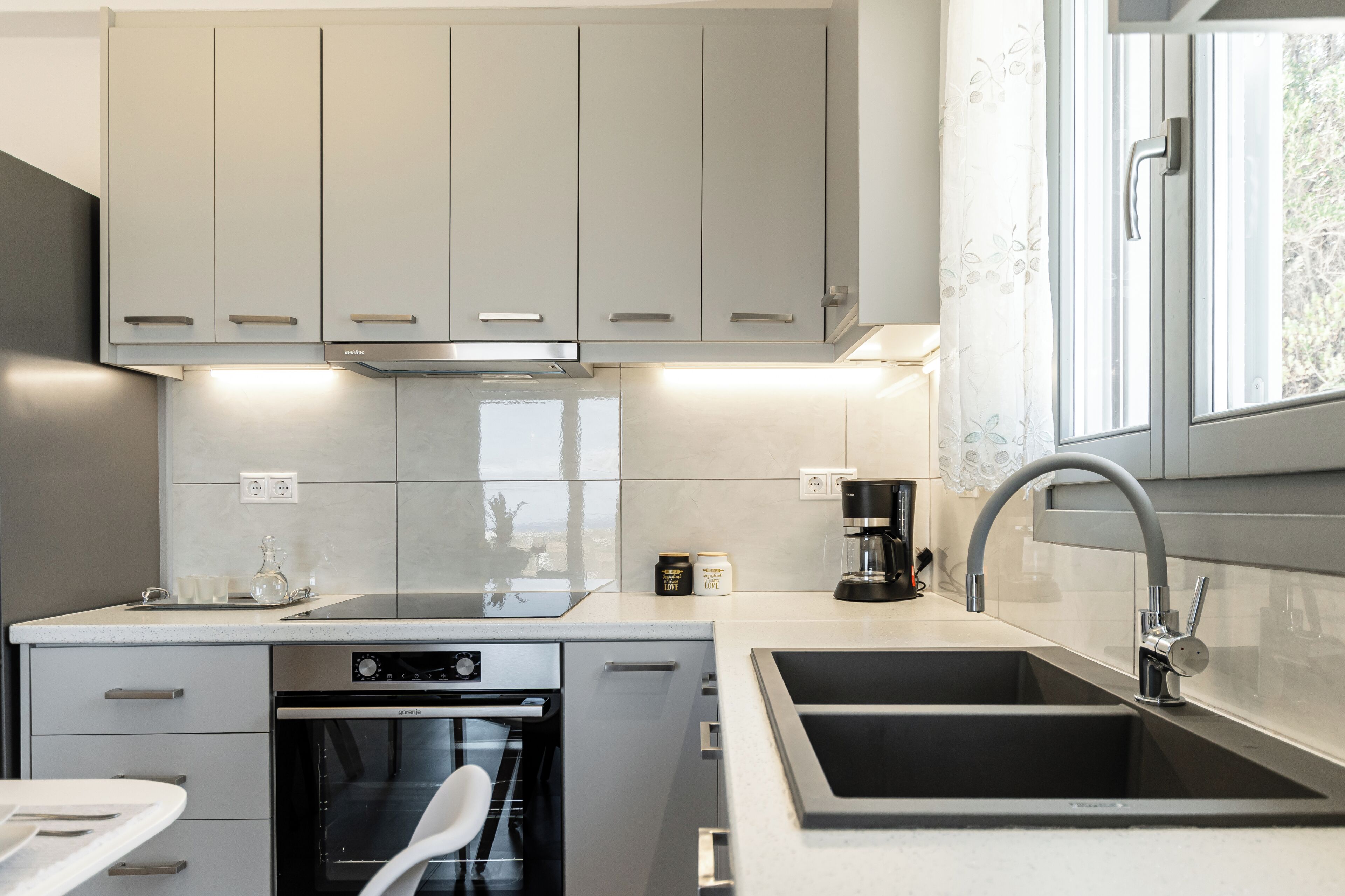 A modern kitchen with grey cabinets, a built-in oven, a coffee maker, and a sink.