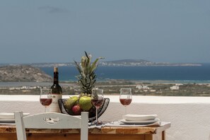 Taula preparada amb ampolla, fruites i gots, amb vista a un paisatge costaner.