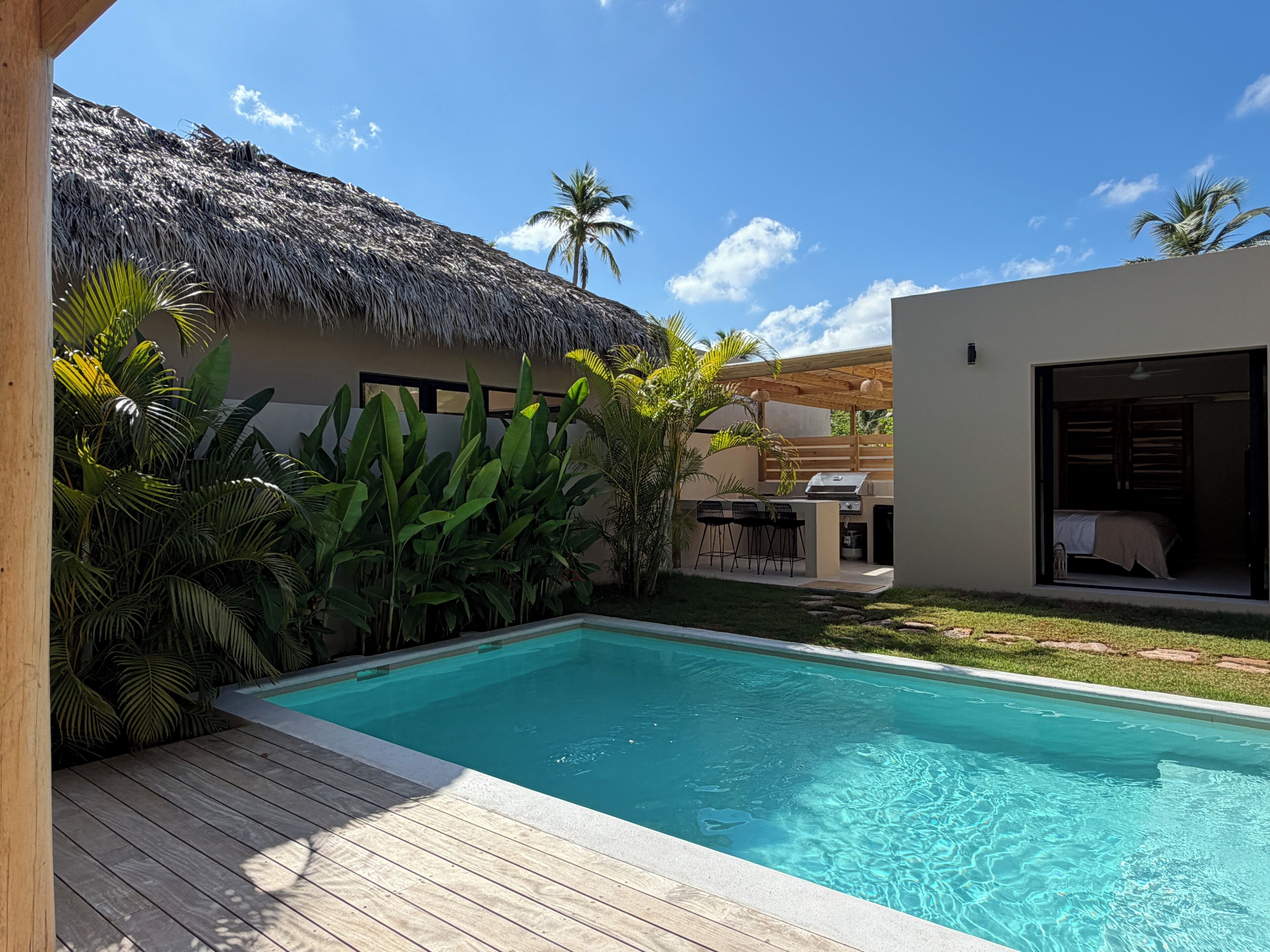 A swimming pool with a wooden deck, a thatched-roof structure, and lush tropical plants.